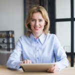 A woman in a blue shirt sits at a table, focused on a tablet in front of her.