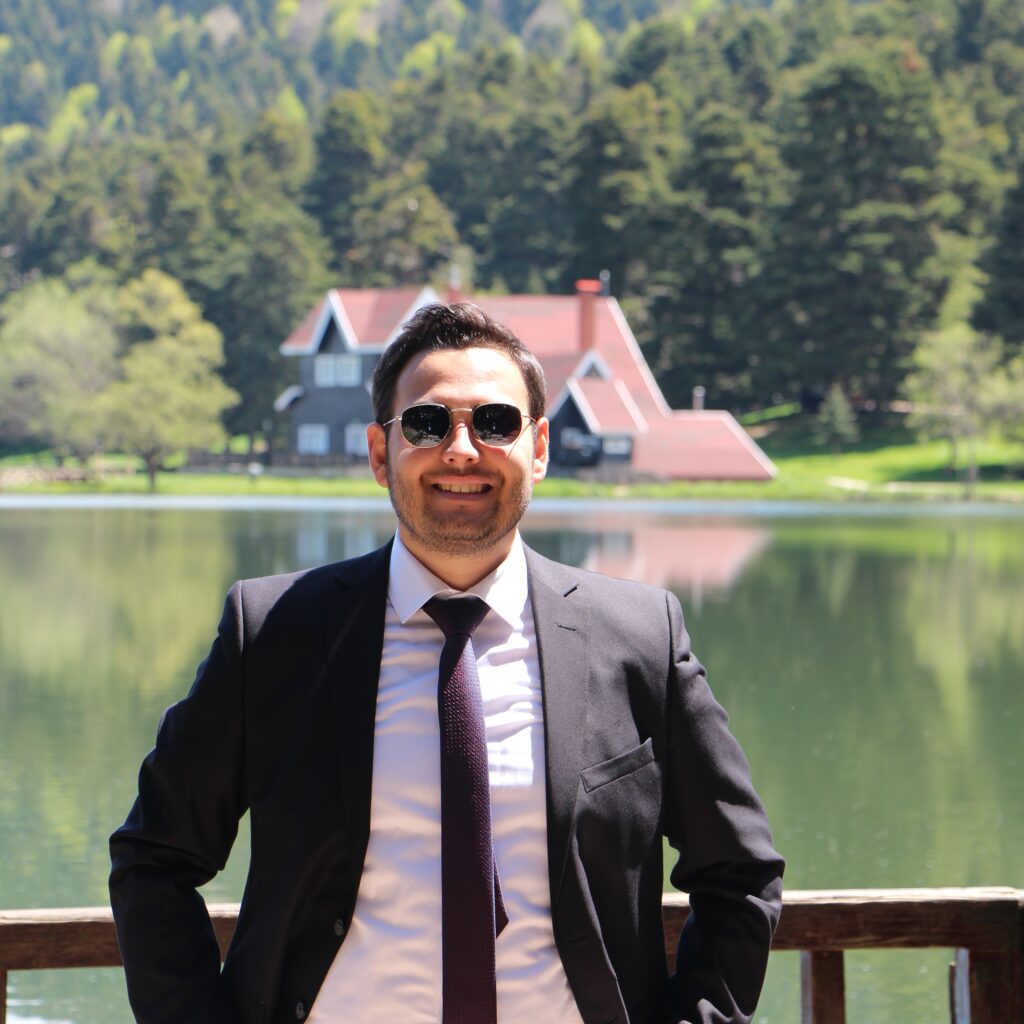 A man wearing a suit and sunglasses poses next to a tranquil lake, surrounded by greenery.